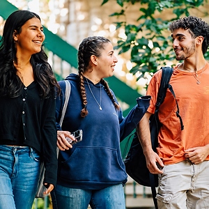 Three young adults with backpacks walk together outdoors, smiling and talking on a campus-like path.